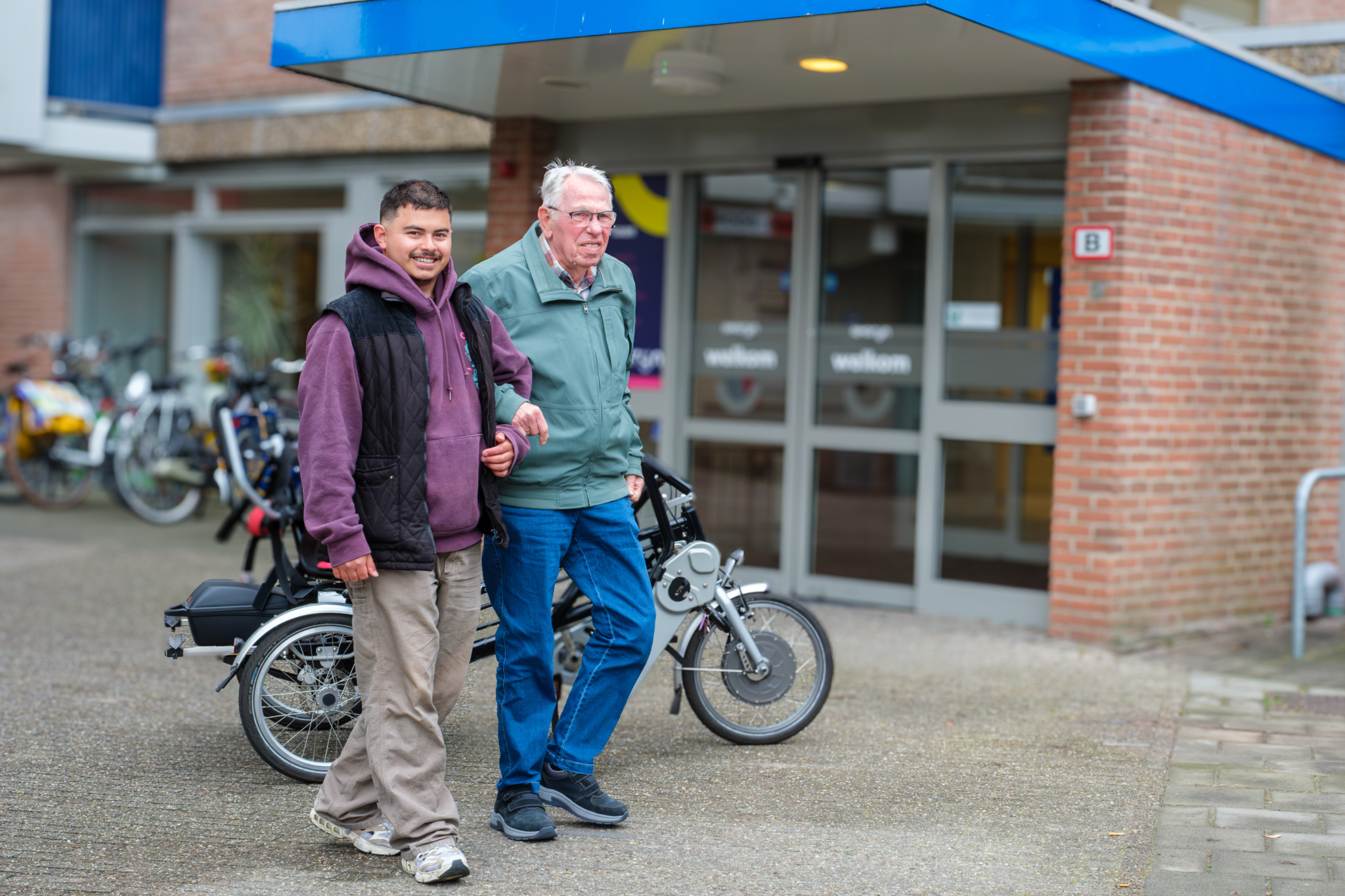Twee mannen lopen samen buiten een gebouw met fietsen op de achtergrond.